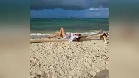 Relaxed woman in a white summer outfit lying on a driftwood log at a sunny tropical beach.
