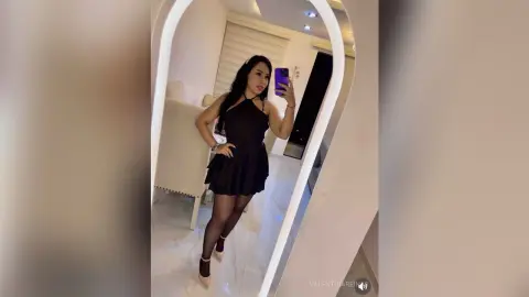 Elegant woman in a black halter mini dress taking a mirror selfie in a modern bright room.