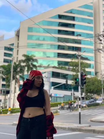 Young woman in a black crop top and red flannel shirt standing on a city street by a modern glass building.