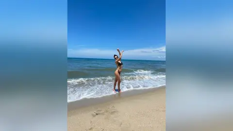 Cheerful woman in a black bikini posing on a sunny beach with ocean waves under a clear blue sky.