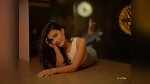 Young woman in a white tank top lying on a wooden floor in a moody, dimly lit indoor setting.