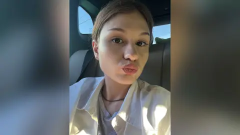 Young woman in a white button-down shirt making a playful pouty face while sitting in a car seat.