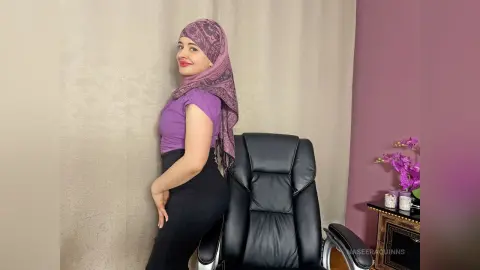Smiling woman in a purple t-shirt and hijab posing in a bright room next to a black office chair.