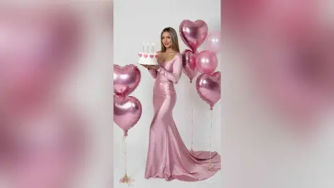 Woman in pink satin evening gown holding a birthday cake surrounded by heart-shaped pink balloons.