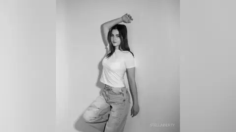 Young woman in white t-shirt and denim jeans posing against a minimalist wall in a moody black and white portrait.