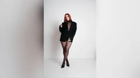 AryaSin poses in a black blazer and sheer black tights against a white background, showing her red hair and legs.