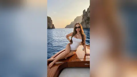 KylieVelvet poses naked on a wooden boat deck, wearing sunglasses and a white bodysuit, with ocean waves and cliffs in the background.