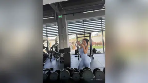 NicoleBella poses in a light blue bodysuit in a gym setting, standing next to a weight rack and dumbbells.