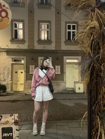 Young woman in a pink hoodie and white skirt taking a selfie on a cobblestone city street at night.