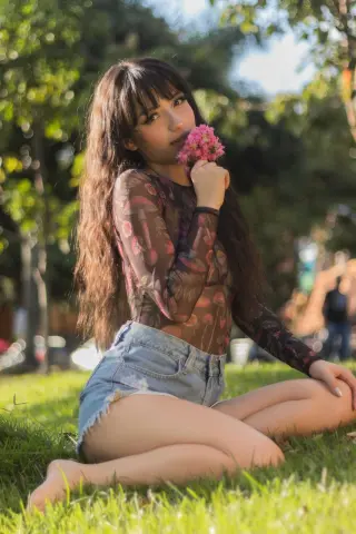 Young woman in a sheer floral top and denim shorts sitting on green grass holding pink flowers in a sunny park.