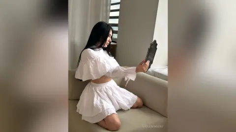 TessaWinsent — TessaWinsent sits on a beige sofa wearing a white crop top and a short white skirt while holding a small mirror.