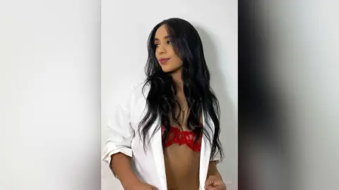 Young woman with long wavy hair wearing a white button-down shirt and red lace bra in a bright minimalist setting.