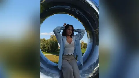 Young woman in a light blue shirt and beige trousers posing inside a large silver ring sculpture on a sunny day.