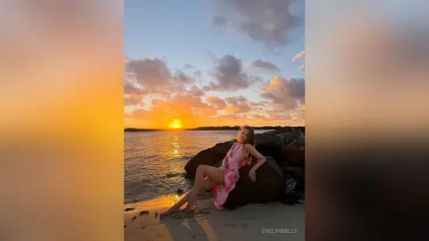 Woman in a pink summer dress sitting on rocks by the ocean during a golden sunset.