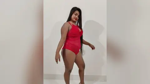 Confident woman in a red lace bodysuit posing against a white wall in a minimalist indoor setting.