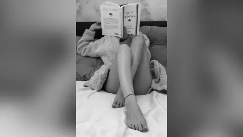 Relaxed woman in a cozy sweater reading an Oprah Winfrey book on a bed in a peaceful, moody black and white setting.