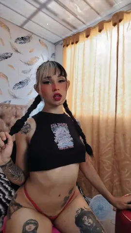 Young woman with blonde bangs and long braids wearing a black graphic crop top in a cozy bedroom setting.
