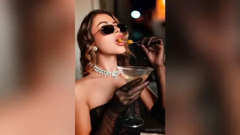 Glamorous woman in black lace and gloves wearing sunglasses and pearls, sipping a martini in a moody lounge.
