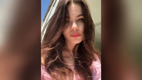 Young woman with long brown hair wearing a pink top smiling in bright natural sunlight.