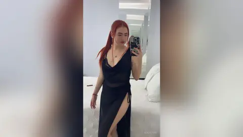 Redheaded woman in a black satin slip dress posing in a bedroom mirror for a moody selfie.