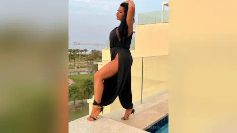 MissyJolie poses on a balcony wearing a black cut-out jumpsuit and black strappy heels.