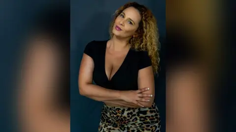 Confident curly-haired woman in a black V-neck top and leopard print skirt posing against a dark studio background.