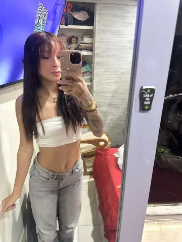 Young woman in a white crop top and grey jeans taking a mirror selfie in a cozy bedroom setting.