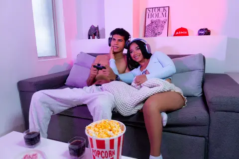 Young couple in loungewear relaxing on a sofa with popcorn and drinks in a cozy, pink-lit room.