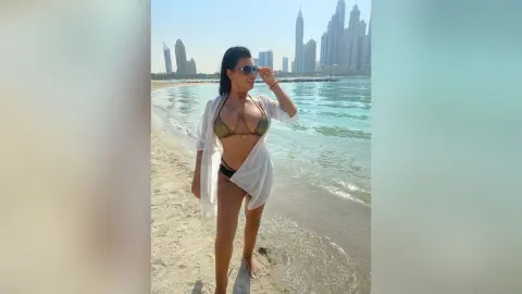 MissyJolie stands on a sandy beach wearing a gold bikini and an open white cover-up shirt with sunglasses.