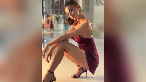 AnaMathers poses in a maroon slip dress, leaning forward with her legs bent and hands resting on her thighs.