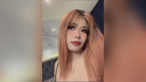 ThaiiTwix looks at the camera with long, light brown hair and red lipstick, wearing a thin black strap against a marble wall background.
