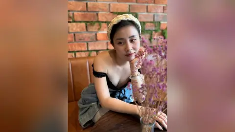 Young woman in a black off-the-shoulder top and headband posing with purple flowers in a rustic cafe.