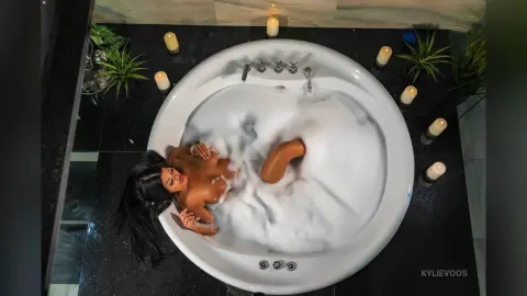 Relaxed woman relaxing in a white bubble bath surrounded by glowing candles in a dark, moody bathroom setting.