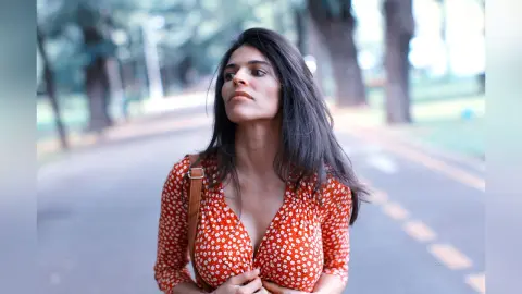 Thoughtful woman in a red floral print blouse walking through a sunlit park setting.