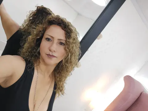 Smiling woman with curly blonde hair wearing a black tank top in a bright, modern indoor setting.
