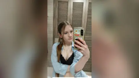 Young woman in a light blue button-down shirt and black lace bra taking a casual mirror selfie at home.