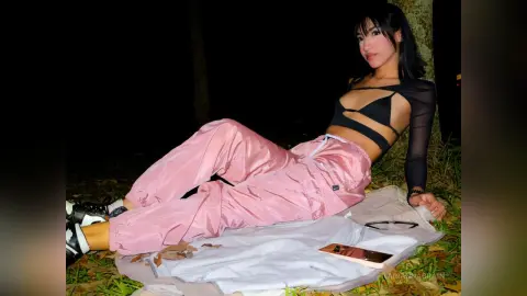 Young woman in pink satin joggers and black crop top relaxing on a white blanket outdoors at night.