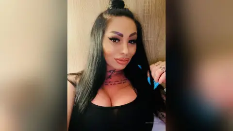 DeeaAnderson poses with long black hair and blue nails, wearing a black tank top that shows her cleavage and neck tattoos.