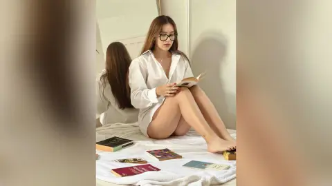 Young woman in white button-down shirt reading a book on a bed in a bright, cozy bedroom setting.