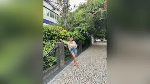 Young woman in a white crop top and denim shorts standing on a cobblestone path near lush green trees.
