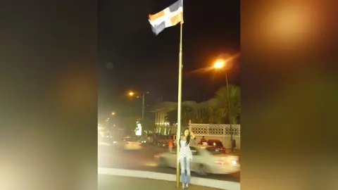 Young woman in a white top standing on a city street at night under a flagpole and streetlights.