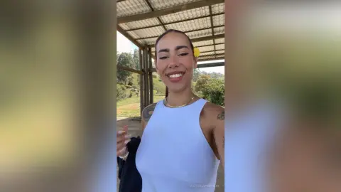 VictoriaFontaine smiles wearing a white tank top and a yellow flower in her hair while standing outdoors in a rural setting.