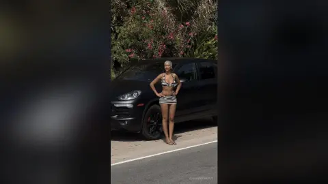 Confident woman in a black and white striped bikini posing next to a black SUV in a sunny outdoor setting.
