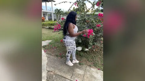 KattyLeya — KattyLeya stands in a garden wearing a white crop top and patterned leggings while touching pink flowers.