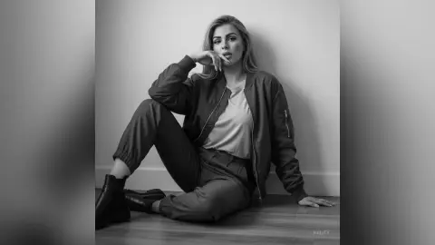 Hailey poses sitting on the floor in a dark bomber jacket and matching pants, looking at the camera with her hand near her mouth.