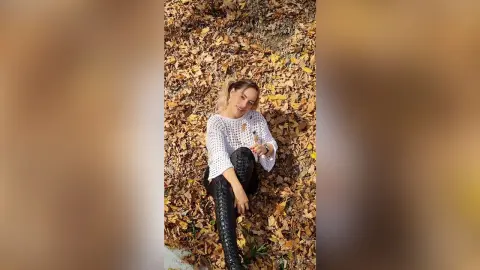 Smiling woman in a white knit sweater and black boots sitting on autumn leaves in a forest.