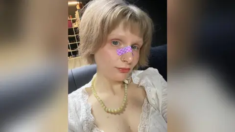 AlitaPatti poses with short blonde hair, wearing a white lace top and a yellow bead necklace, showing cleavage and a purple scale-patterned patch on her nose.