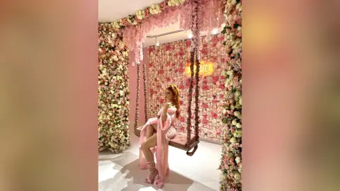 Elegant woman in a pink silk gown sitting on a floral swing in a dreamy rose-filled room.