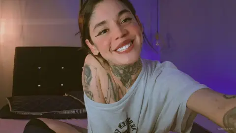 Smiling woman with neck tattoos wearing a white t-shirt in a dimly lit bedroom setting.