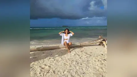 Young woman in white summer outfit sitting on a driftwood log at a serene tropical beach under a cloudy sky.